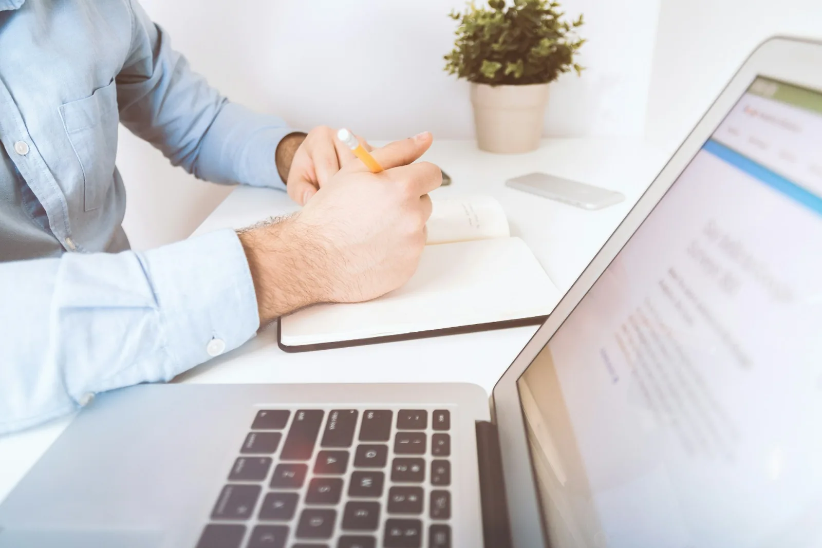 Write a Press Release That: Turn Inquiries Into Enrolled image A person in a light blue shirt holding a pencil and writing in a notebook at a white desk, with an open laptop.