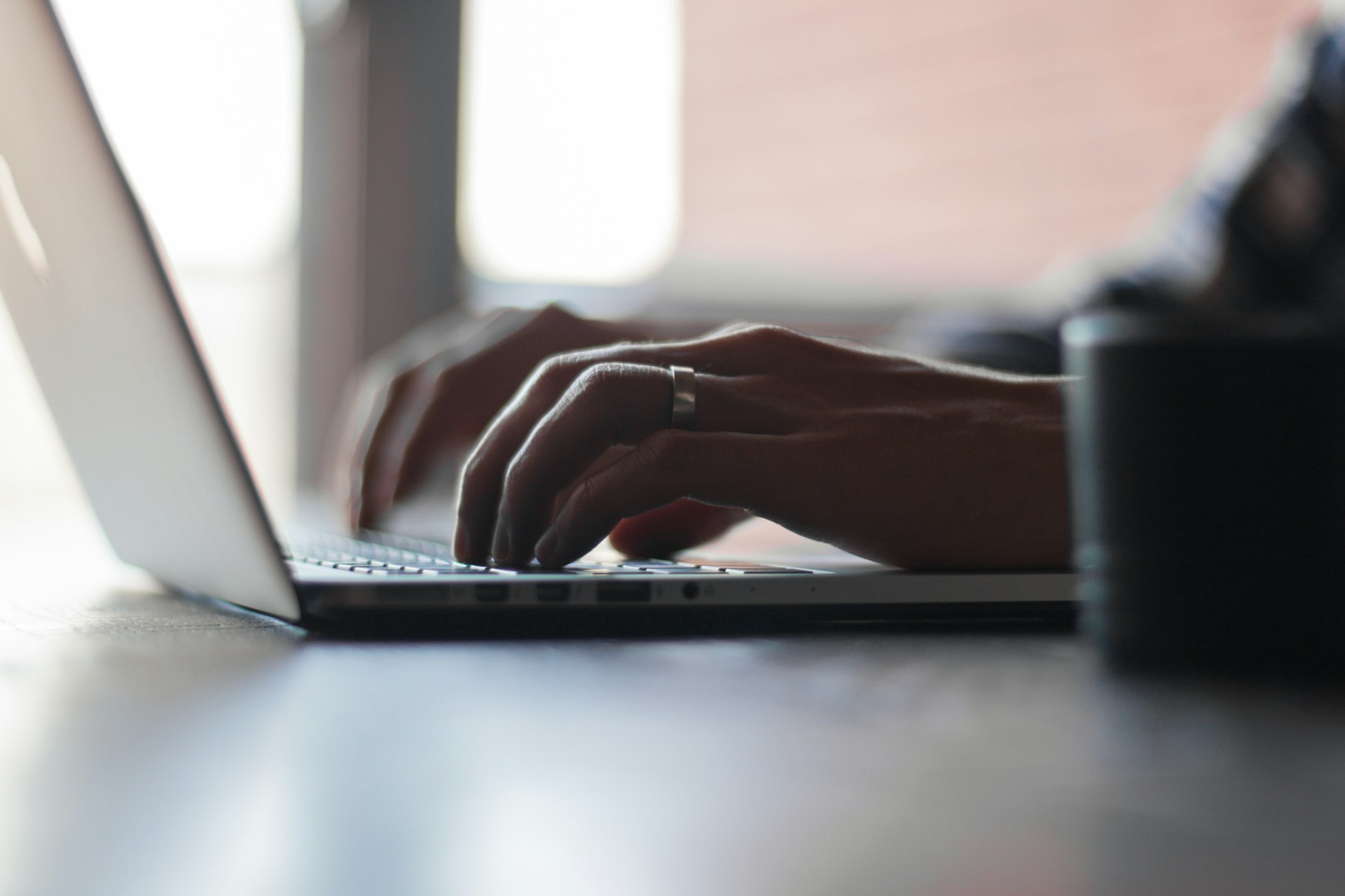 Write a Press Release That: Turn Inquiries Into Enrolled image A person typing on a laptop keyboard with both hands in a softly lit workspace with a blurred coffee mug in the foreground.