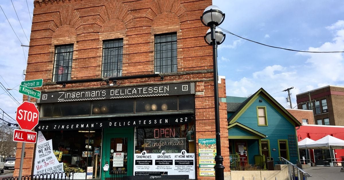 Exterior photo of Zingerman&rsquo;s Delicatessen in Ann Arbor, a brick corner storefront with a &lsquo;Zingerman&rsquo;s Delicatessen&rsquo; sign, street signs (Detroit St and E Kingsley St), a stop sign, and an &lsquo;OPEN&rsquo; window sign.